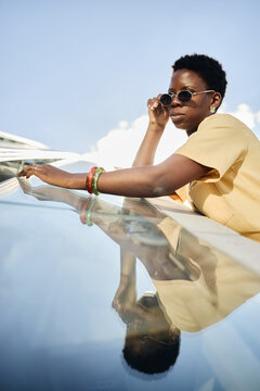 Vertical shot of young Black woman adjusting sunglasses while leaning on glass panel, reflecting confident travel lifestyle and personal freedom. Ideal for mobility, summer campaign