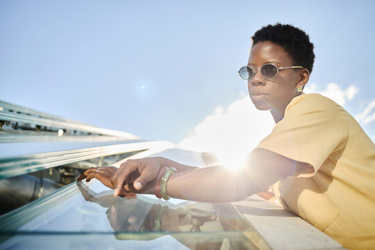 Young adult Black woman leaning on glass railing outdoors, wearing sunglasses and looking away, conveying urban confidence and modern lifestyle. Ideal for fashion, travel, independence marketing