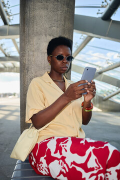 Vertical shot of young Black woman using smartphone while sitting in transit hub, managing mobile communication and travel planning. Ideal for digital lifestyle or mobility marketing