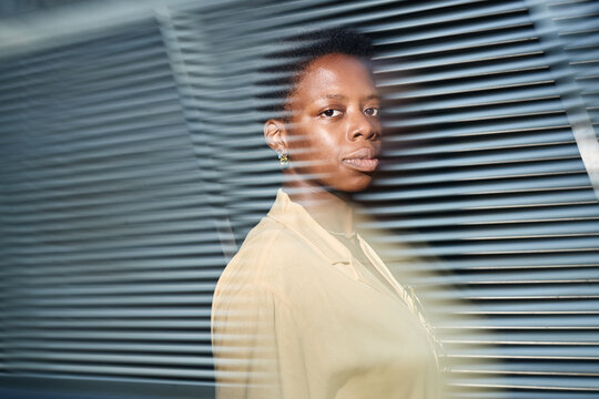 Portrait of young adult Black woman standing behind window blinds, looking at camera and conveying privacy, focus, corporate identity. Ideal for workplace branding or leadership campaigns