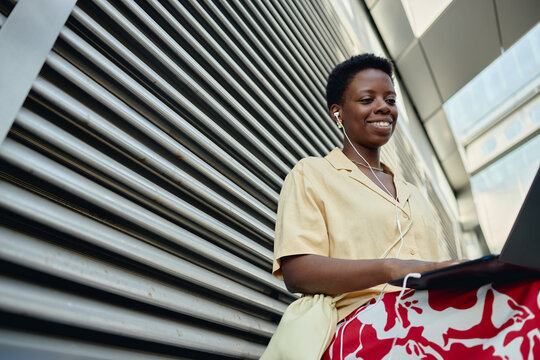 Young adult Black woman using laptop and earphones while sitting outdoors, working remotely and smiling. Ideal for remote work, digital nomad, mobile productivity marketing