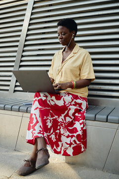 Vertical shot of young adult Black woman working on laptop while listening through earphones, sitting outdoors and managing mobile workflow. Useful for remote work, freelance, digital nomad marketing