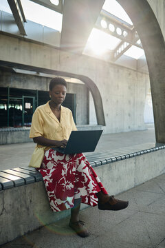 Vertical shot of young Black woman working on laptop while sitting outdoors in urban transit setting, representing mobile productivity and remote work. Ideal for hybrid work campaigns