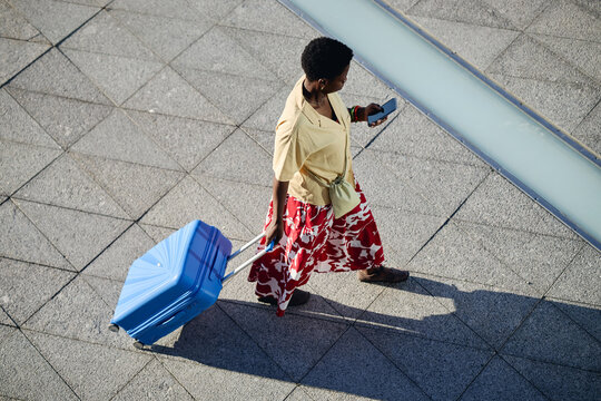 High angle view of young adult Black woman pulling wheeled suitcase while checking smartphone during travel, representing mobile lifestyle. Ideal for tourism, business trip, app promotion