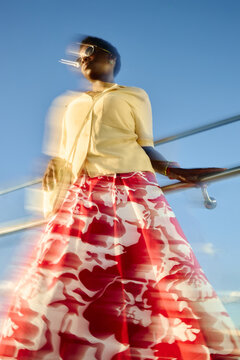 Vertical shot of Black young adult woman wearing colorful skirt and sunglasses standing outdoors in motion blur, expressing urban confidence. Ideal for fashion marketing, lifestyle branding