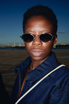 Vertical portrait of young adult African American woman wearing sunglasses, facing camera outdoors near waterfront, conveying confidence. For fashion branding, lifestyle ads, identity campaigns