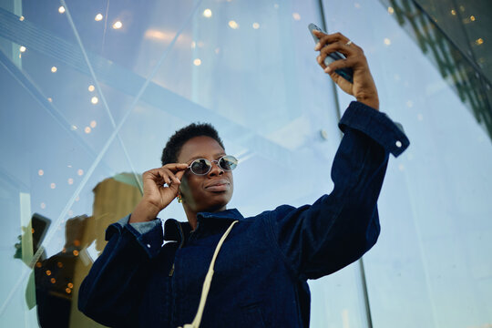 Low angle shot of Black young adult woman holding smartphone and adjusting sunglasses while taking selfie outdoors, conveying mobile lifestyle and personal branding for social media marketing