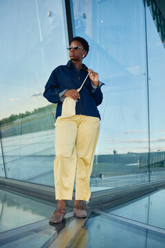 Vertical shot of young adult Black woman standing by glass facade, holding handbag and looking away in urban setting. Suitable for fashion marketing, lifestyle branding