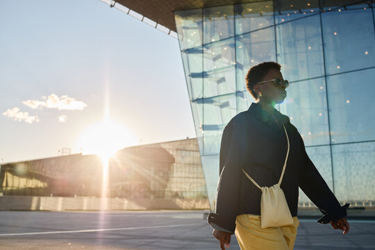 Medium shot of African American young adult woman walking past modern office building, wearing sunglasses and carrying bag, conveying urban business mobility. For corporate branding or career content