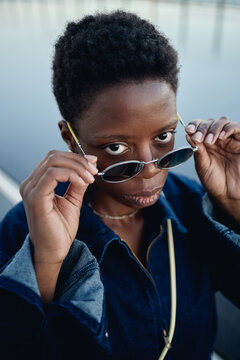 Vertical portrait of young Black woman lowering sunglasses and looking at camera, conveying confidence and urban style. Ideal for fashion marketing, personal branding, lifestyle campaigns