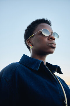 Vertical shot of young Black woman wearing sunglasses, looking away against open sky, conveying confidence and modern identity. Useful for fashion branding, lifestyle campaigns