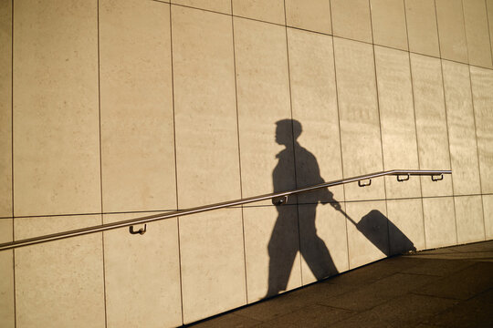 Traveler shadow pulling rolling suitcase along urban wall, suggesting business trip, commute, or transit movement. Wide shot, useful for travel marketing, mobility services, tourism ads