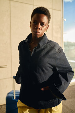 Vertical portrait of young adult Black woman standing with suitcase and looking at camera, conveying business travel and mobility. Useful for corporate travel, airport service, relocation marketing