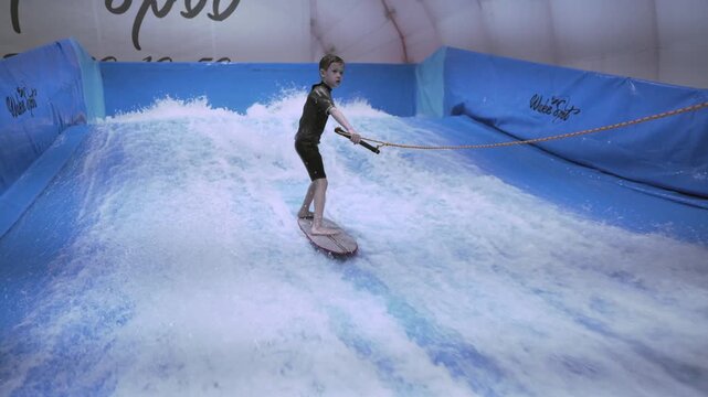 Teenager is riding surf board on wave simulator indoors. Young surfer during training on generated waves. Water sport activity. Surfer kid enjoy surfing on indoor surf imitator. Surfing training.
