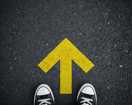 First person view of feet in sneakers standing before a painted yellow arrow on black asphalt pointing forward direction with direction pavement sneakers decision navigation opportunity