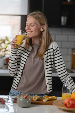 Smiling young woman enjoying a glass of fresh orange juice during a healthy breakfast with chia pudding and fruit in a modern kitchen, promoting wellness and balanced nutrition