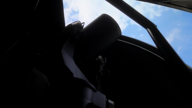 A low angle shot from inside a dark observatory dome showing the silhouette of a large professional telescope pointing upwards through the open slit towards a bright blue sky.