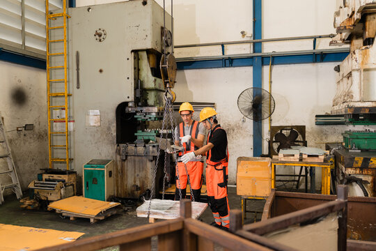 Two engineers review a laptop beneath a chain hoist load, flanked by two old power press machines on the factory floor