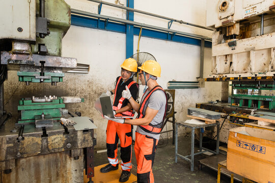 Two engineers stand between a pair of metal stamping presses, one giving a thumbs-up, holding a laptop, on the factory floor