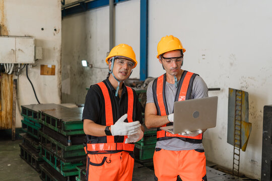 Engineers in orange vests discussing production data on laptop among stacked steel dies indoors