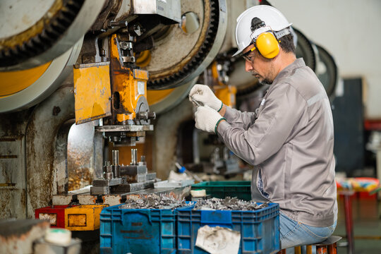 Machine operator examining small stamped metal component beside mechanical power press