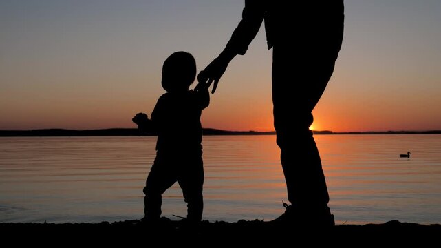 Silhouetted mother holding her small toddler hands, teaching baby to walk on beach during golden sunset. Calm water reflecting the glowing sun on the horizon. The first steps of a child