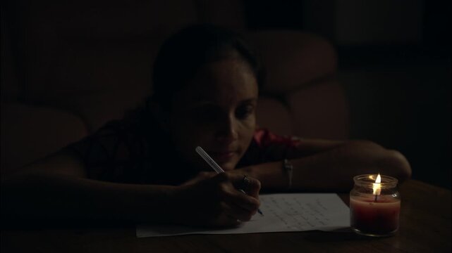 A moody low-light photograph of a woman writing on paper beside a burning candle in a quiet room.