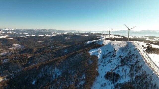 Aerial view of wind turbines on a snow-covered mountain range under a clear blue sky. Drone footage