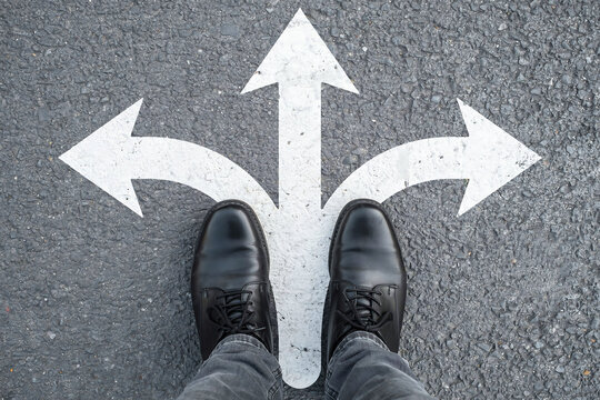 Professional standing at three way intersection looking down at directional arrows with crossroads decision direction business strategy management planning thinking motivation leadership