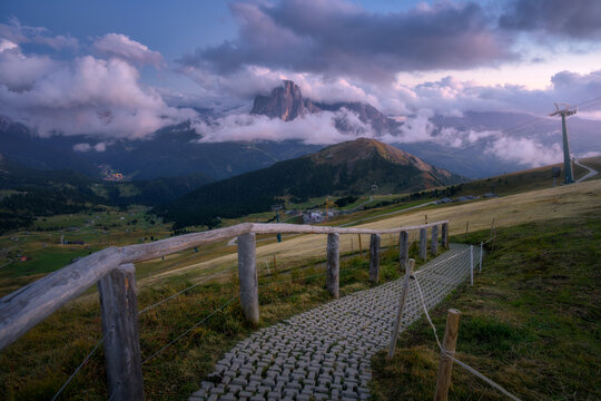 Paved walking path with wooden fence and mountains in fog at sunset. Dolomites, Italy. Mountain trail, green valley, alpine meadows, purple sky, clouds and rocky peak at dusk in summer. Landscape