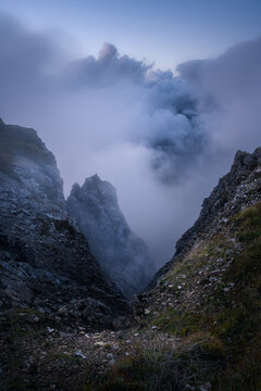 Dramatic mountain gorge with rocky cliffs surrounded by thick clouds and fog at dusk. Dolomites, Italy. High altitude alpine canyon landscape at sunset with fog rising over steep mountain rock slopes