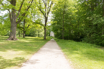Obraz premium A scenic landscape view of historic Herttoniemi Manor Park in Helsinki, Finland, featuring sun-drenched paths, majestic old deciduous trees, and lush green lawns under a bright summer sky.