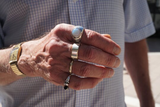 Schlanke Hand von &auml;lterem stehendem Mann mit Silberringen an allen Fingern vor Oberk&ouml;rper mit blau-wei&szlig;em Karohemd in Stadt bei Sonne am Morgen im Sommer
