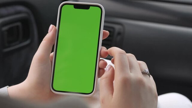 Person holds phone with green screen inside car. Fingers tap screen. Ring visible on finger. Ideal for app mockups or driving-related ads