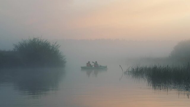 Couple enjoying journey paddling boat across tranquil lake enveloped in dense morning fog during a beautiful, serene sunrise with warm, glowing light. Romantic date of a loving couple on a boat trip