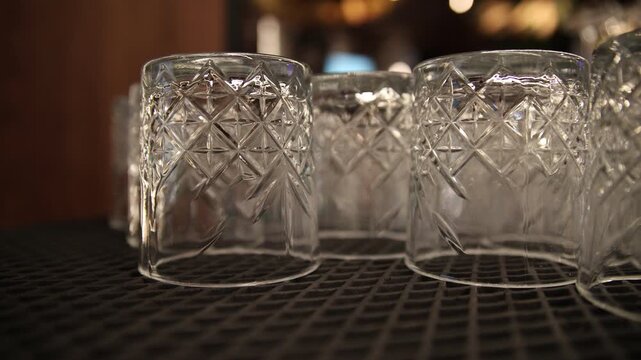 Empty inverted crystal cut-glass tumbler glasses resting on a black rubber bar mat surface with blurred warm bokeh lights and a cozy nightlife pub restaurant interior atmosphere.