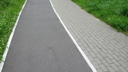 Obraz premium Green urban park pathway combining asphalt walking trail and paved sidewalk surrounded by grass and natural landscape