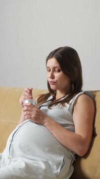 Pregnant woman relaxing on a comfortable sofa at home, eating delicious yogurt from a small container with a spoon, embracing a healthy lifestyle and proper nutrition during her pregnancy