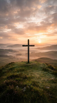 Wooden cross standing on misty hill at sunrise. Solitary religious symbol on green grassy peak during golden hour.