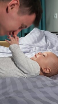Young, caring father gently playing with his adorable infant son or daughter lying on a bed. The happy baby smiles and touches his dad's face, showcasing a beautiful bond of love and family
