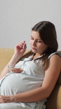 Pregnant woman resting on the couch, thoughtfully eating dates from a glass bowl that is ingeniously balanced on her baby bump, highlighting a healthy diet during maternity