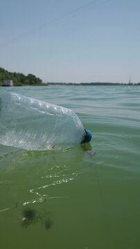 An empty clear plastic bottle floats vertically in the green water of a lake under a blue sky. Concept of environmental pollution, water contamination, and ecological conservation.
