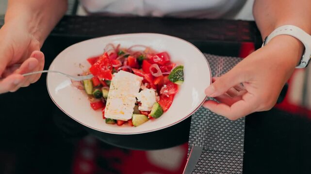 POV of hands using a fork to eat fresh greek salad with feta cheese outdoors.