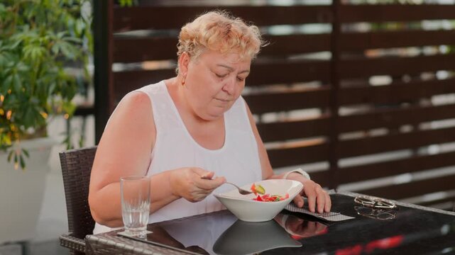 Senior overweight woman eating fresh vegetable salad at outdoor restaurant.