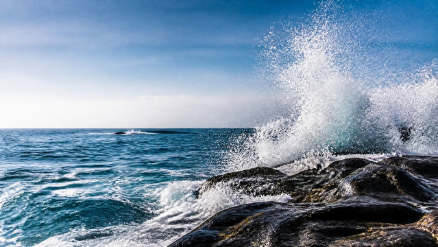 Dynamic ocean wave crashing against rugged dark coastal rocks under a bright blue sky, representing power, nature, freedom, and resilience, perfect for travel, environmental, or lifestyle