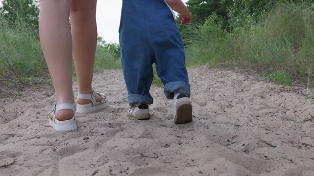 A mother and her young toddler child walk together down a sandy forest path seen from behind. Concept of early childhood development, family bonding, parenting, and outdoor growth.