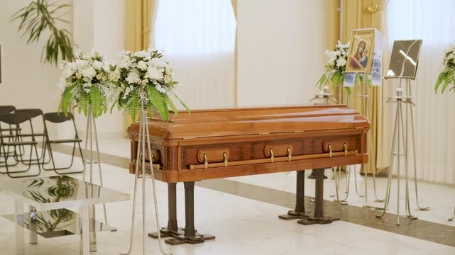 Closed wooden coffin in a bright funeral home or chapel. Solemn farewell ceremony setup with white floral arrangements, empty chairs, and a religious icon.
