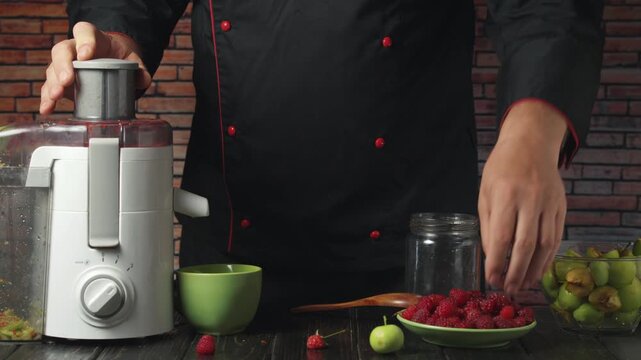 Chef in black uniform prepares fresh raspberry juice using a juicer, placing raspberries into the machine while surrounded by green fruits and jars on a wooden kitchen table