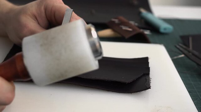 Craftsman perforating dark leather with mallet and chisel tool for stitching handcrafted accessory in a workshop