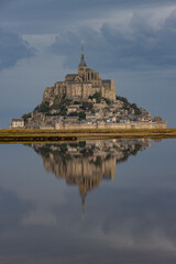 Fototapeta premium Atmospheric vertical shot of Mont Saint-Michel casting a pristine mirror reflection onto calm tidal waters under a dark stormy sky in Normandy.